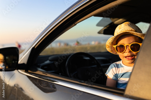 Fashionable baby boy, peeks out of the car in the sunset. Beach and sea in the background. Travel summer vacation concept. © zphoto83