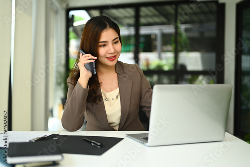 Gorgeous millennial female entrepreneur having phone conversation and using laptop at desk