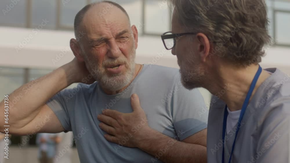 Health care specialist sits with aged patient on the bench and discusses his treatment. Elderly man talks with doctor and complains of neck pain. Mature professional in medical uniform works outdoor. Health care specialist sits with aged patient on the bench and discusses his treatment. Elderly man talks with doctor and complains of neck pain. Mature professional in medical uniform works outdoor.