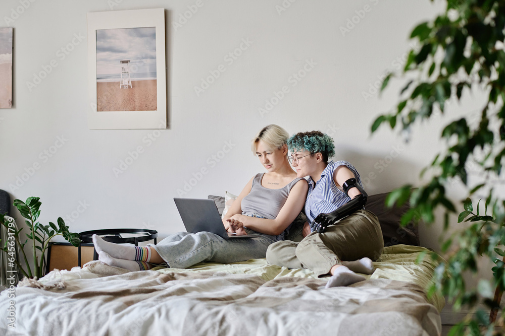 Young lesbian couple relaxing together on bed at home and surfing the net on laptop Young lesbian couple relaxing together on bed at home and surfing the net on laptop