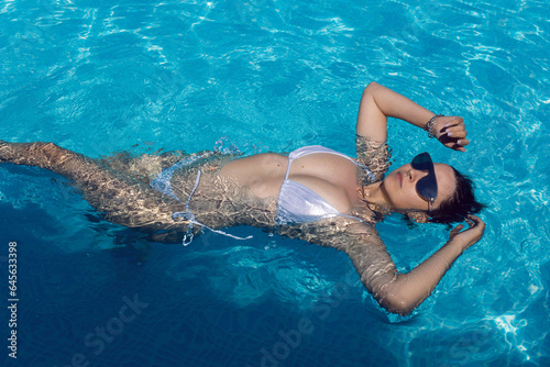 Fashion stylish a sexy woman in a silver swimsuit bikini and sunglasses is lying on her back in a blue pool in summer under the sun outdoors.
