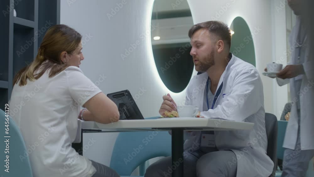 Medics rest in hospital cafeteria, have meal and talk. African doctor brings coffee and sits down to colleagues. Caucasian doctor watches tests results on digital tablet, discusses with other medics. Medics rest in hospital cafeteria, have meal and talk. African doctor brings coffee and sits down to colleagues. Caucasian doctor watches tests results on digital tablet, discusses with other medics.