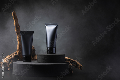Black cosmetic squeeze tubes on black circular podium decorated with wood log on black background with smoke.