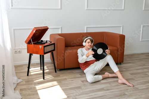 Teenager boy holding vinyl record in hands, sitting on floor near turntable and sofa. Minimalist home interior, lifestyle
