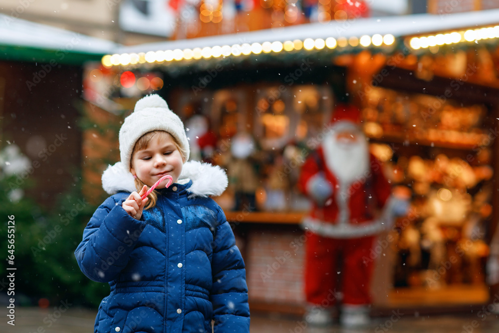 Little cute preschool girl with candy cane from a sweets stand on Christmas market. Happy child on traditional family market in Germany. Preschooler in colorful winter clothes during snowfall