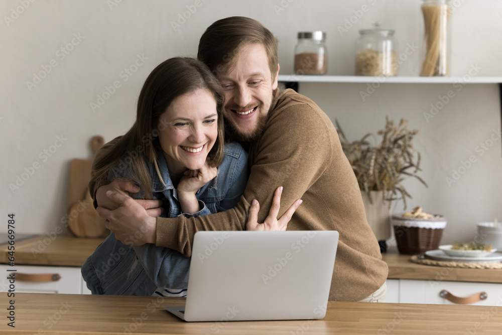 Positive overjoyed young husband and wife hugging at laptop in joy, looking at display, smiling, laughing, getting excited, happy, celebrating luck, good news, success, achievement, win Positive overjoyed young husband and wife hugging at laptop in joy, looking at display, smiling, laughing, getting excited, happy, celebrating luck, good news, success, achievement, win