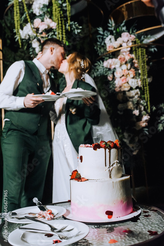 Stylish happy groom and beautiful young bride hugging, kissing while standing in a decorated arch at night holding a piece of cake on a plate. Wedding photography, portrait.