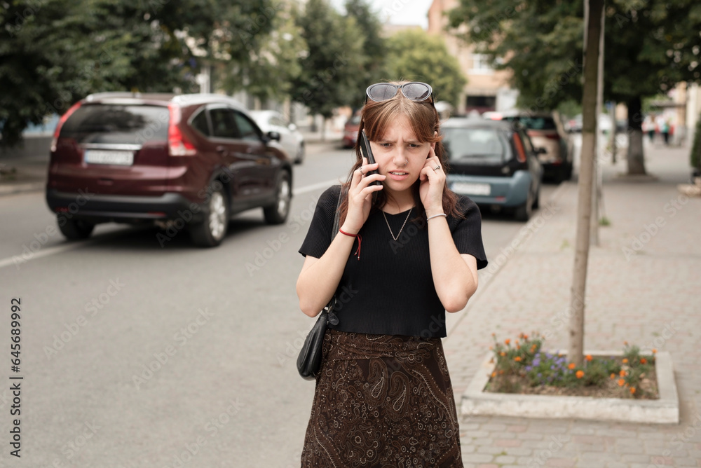 A girl is talking on the phone in the city and covers one ear, she has a very important conversation, but because of the noise of the cars, she cannot hear the interlocutor well