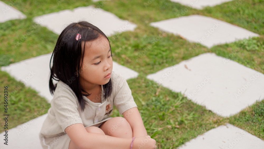 Asian little girl sitting pensively on the grass, hugging her knees. Asian little girl sitting pensively on the grass, hugging her knees.