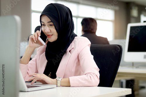 Beautiful Muslim Asian woman officer with hijab, talking with customer by mobile phone while working on laptop computer at office desk with her busy working colleagues. Female worker at workplace.