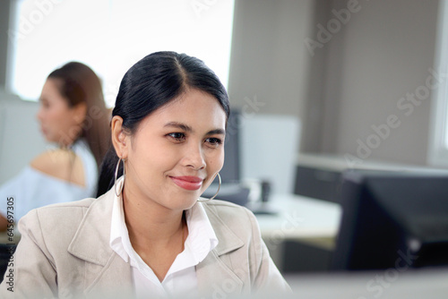 Portrait of happy beautiful Asian woman officer sitting and working at office desk with blurred background of her busy working colleagues. Pretty attractive smiling female staff worker at workplace.