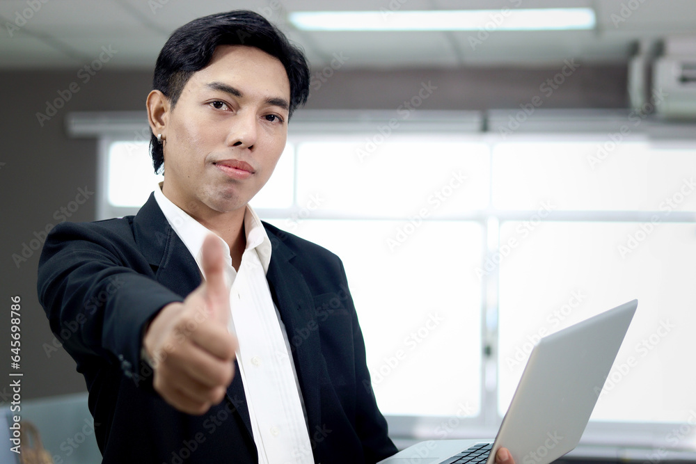 Portrait of happy smiling Asian businessman in black suit giving thumb up to camera while holding laptop computer, standing inside office building, confident male officer working at workplace. Portrait of happy smiling Asian businessman in black suit giving thumb up to camera while holding laptop computer, standing inside office building, confident male officer working at workplace.