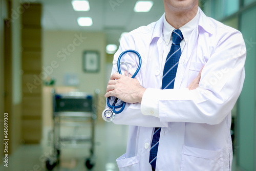 Confident male doctor in white workwear holding stethoscope standing with arms crossed in corridor of medical clinic, doctor working at hospital. medical and health care concept.