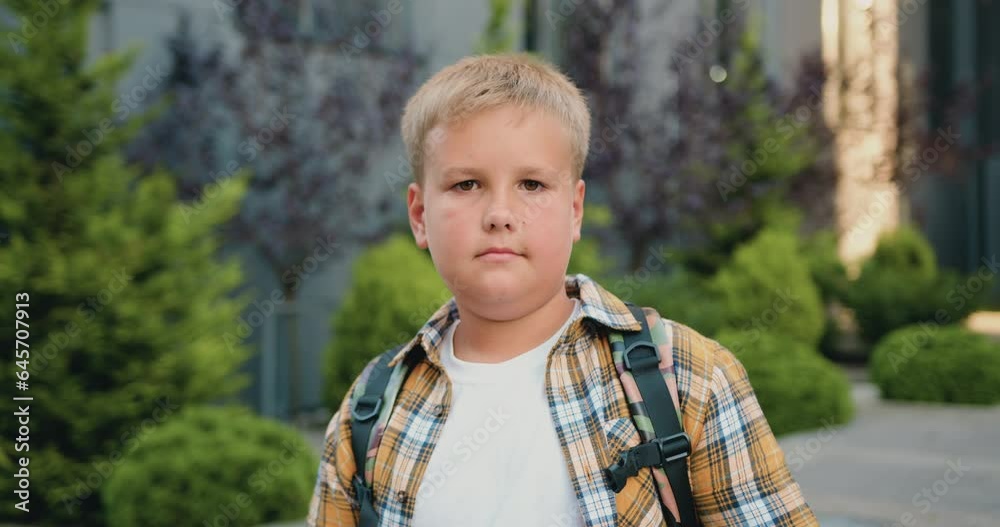 Close up of pensive good- looking caucasian schoolboy with backpack posing and looking at camera in school building background, outdoors. Elementary preteen school student portrait Close up of pensive good- looking caucasian schoolboy with backpack posing and looking at camera in school building background, outdoors. Elementary preteen school student portrait