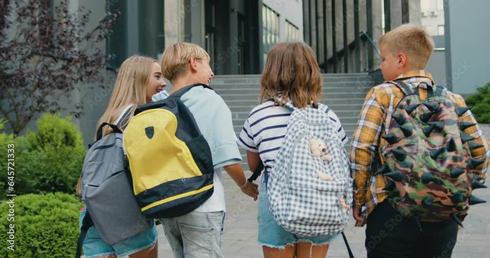 Group of four smart diverse classmates with colorful backpacks are having fun talking while going to school school, outdoors. Back view.