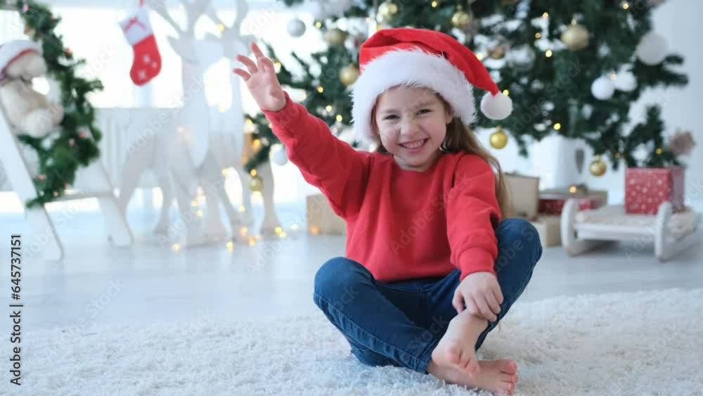 Little child girl smiling and waving hand in Christmas time wearing Santa hat and sitting on fluffy carpet. Pretty kid in decorated New Year home with festive tree