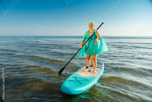 A woman in a turquoise swimsuit with a skirt and a scarf on her head on a SUP board swims to the seashore. © finist_4