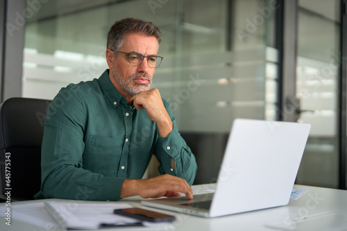 Busy older professional business man entrepreneur looking at laptop working at desk. Serious mid aged executive manager investor using computer thinking on digital financial market analysis in office.