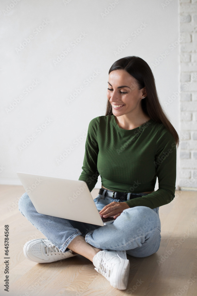 Young cute confident student woman in casual clothes is using a laptop and smiling while sitting on the floor, on gray background. Study, education, university, college, graduate concept Young cute confident student woman in casual clothes is using a laptop and smiling while sitting on the floor, on gray background. Study, education, university, college, graduate concept