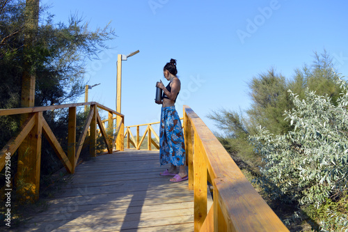 A girl in a long skirt with a jug in her hands walks along a wooden bridge that is by the sea.