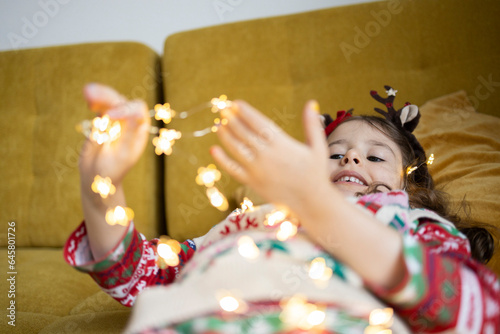 Happy cute girl in Christmas pullover playing with string light at home