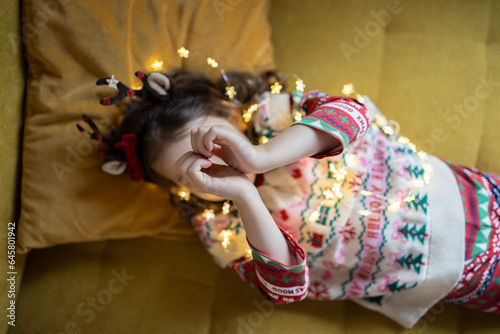 Happy four years girl in Christmas pullover and with Christmas lights shows a heart with her hands 