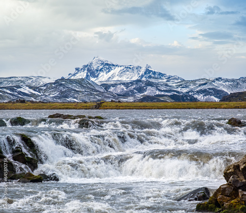 Season changing in southern Highlands of Iceland. Picturesque waterfal Tungnaarfellsfoss panoramic autumn view. Landmannalaugar mountains under snow cover in far.