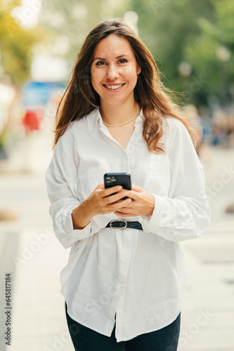 Vertical street portrait of a smiling at the camera woman walking and holding her phone. © Vulp