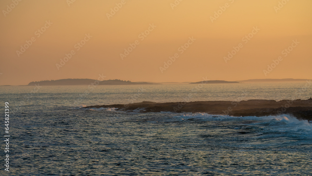 Vibrant coastal sunset paints the sky in hues of orange and purple, casting a warm glow on the rugged rocky shore, a tranquil moment captured in nature's artistry.