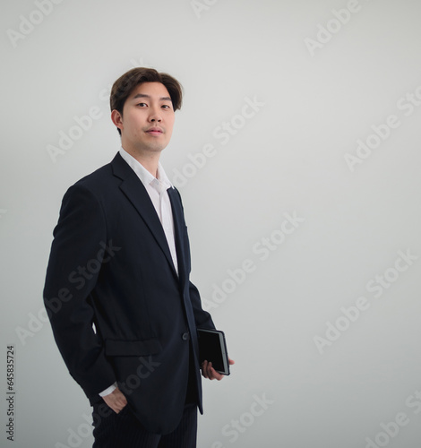 Business man portrait Asian one person wearing a black suit, handsome cool sharp smart standing looking, hand holding mobile phone and laptop ready for work inside the office, with white background