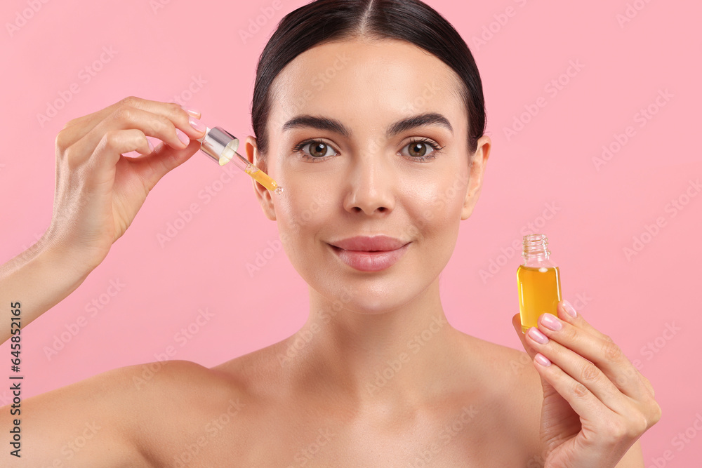 Fototapeta premium Beautiful young woman applying serum onto her face on pink background