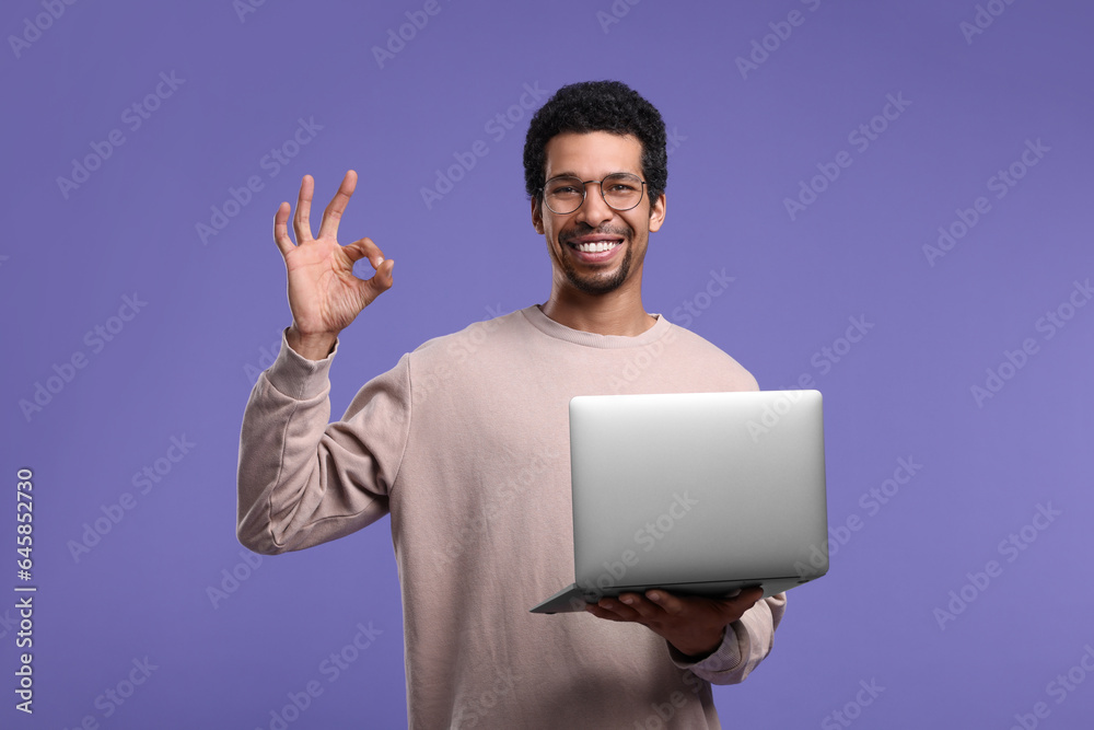 Smiling man with laptop showing ok gesture on purple background Smiling man with laptop showing ok gesture on purple background