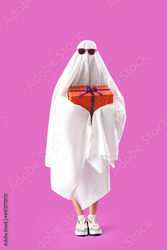 Young woman dressed as ghost with Halloween gift on pink background
