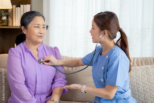 Young asian nurse or caregiver sitting on sofa holding stethoscope listen heartbeat with senior woman in living room at home, doctor check cardiologist disease with elderly, medical and insurance.