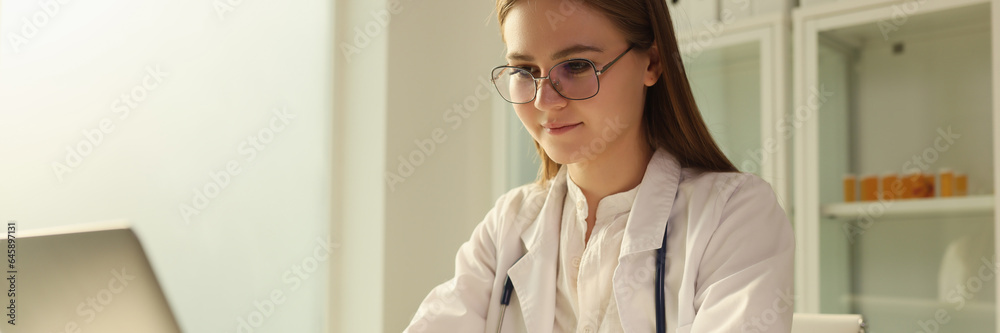 Young female doctor works on laptop computer at table in medical office.