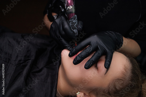 In the cosmetology room, the procedure of permanent lip makeup using a tattoo machine is being carried out. Delicate permanent lip makeup for blondes