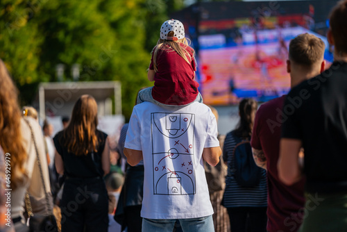 Man holding a kid on a shoulders wearing a baseball cap during public sports event of basketball in summer wearing tshirts photo