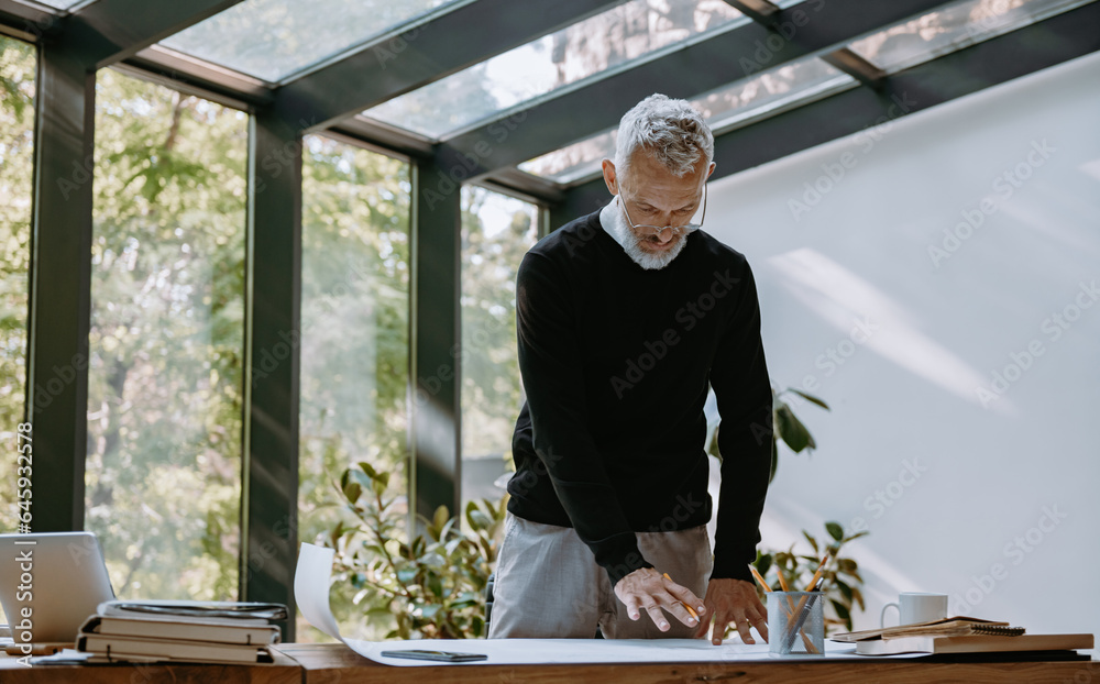 Confident mature architect examining blueprint while working in the creative office Confident mature architect examining blueprint while working in the creative office