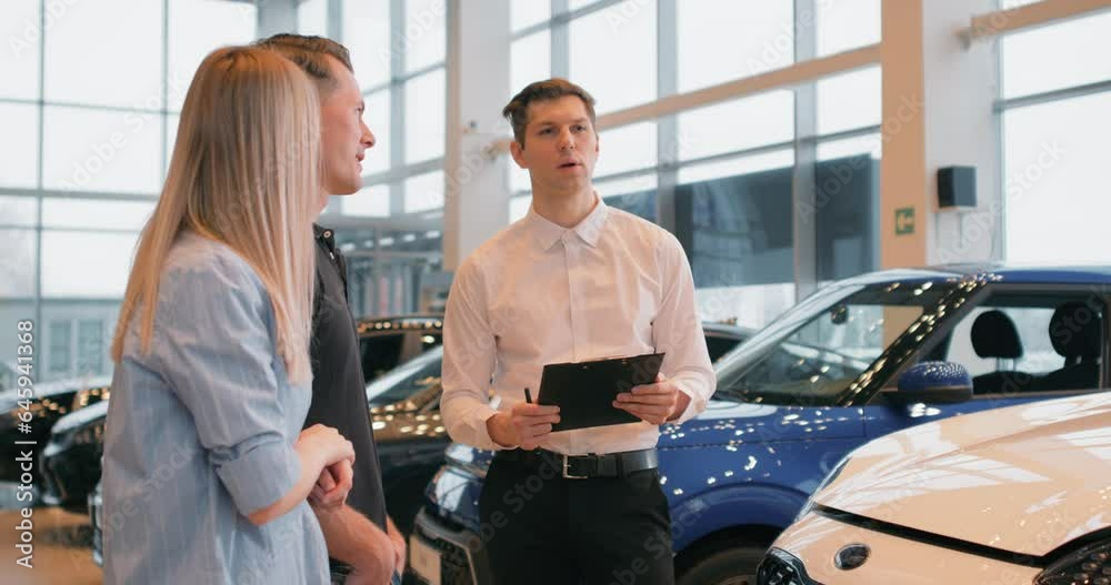 Car Dealership. Cheerful Elegant Manager Guy Showing Automobile To Customers In Auto Dealership Store. Young sales agent helping adult couple to choose a new car in modern car showroom. Car Dealership. Cheerful Elegant Manager Guy Showing Automobile To Customers In Auto Dealership Store. Young sales agent helping adult couple to choose a new car in modern car showroom.