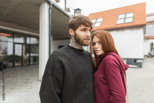 Cool handsome fashionable hipster man with a beard in a black pullover and a beauty redhead stylish model girl in a red sweater are standing together in the city. fashion couple © alones