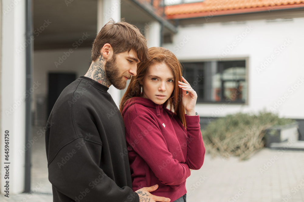 Handsome bearded hipster man hugging beautiful red-haired girl in trendy fashion pullover outdoors. Fashion street couple