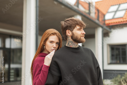 Fashion beautiful young stylish couple bearded hipster man and red-haired beautiful woman in fashionable hoodie on the street