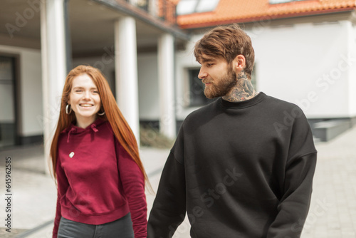 Happy beautiful young fashionable couple hipster man with beard and cute pretty cheerful red-haired girl in fashion outfit with hoodie walking on the street