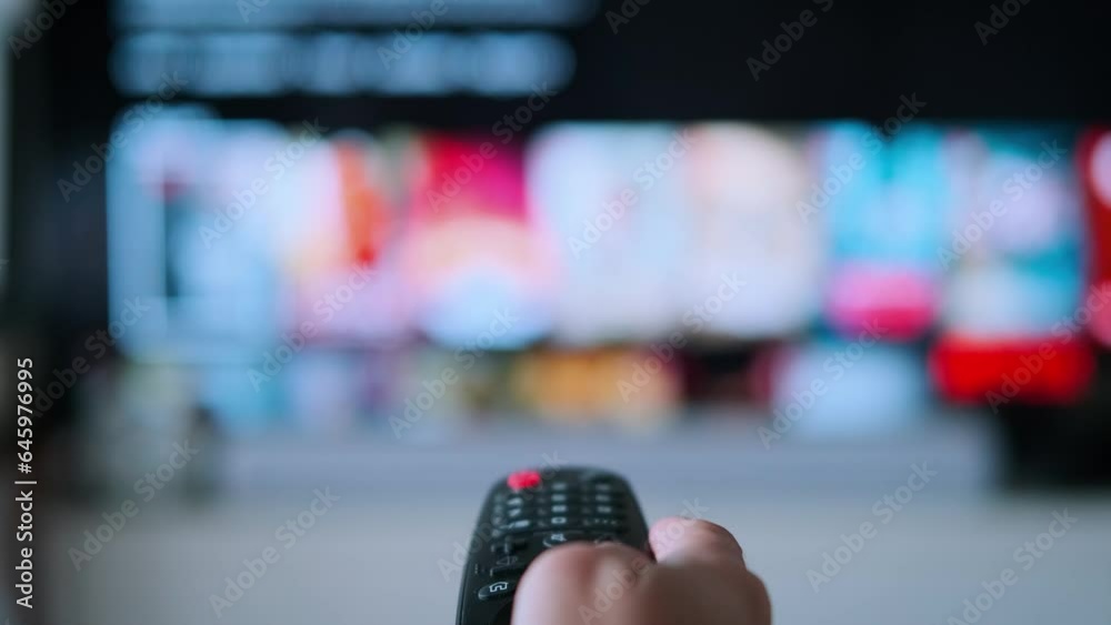 Woman hand selects internet tv channels with remote control. Close up view. Blurry tv at the background