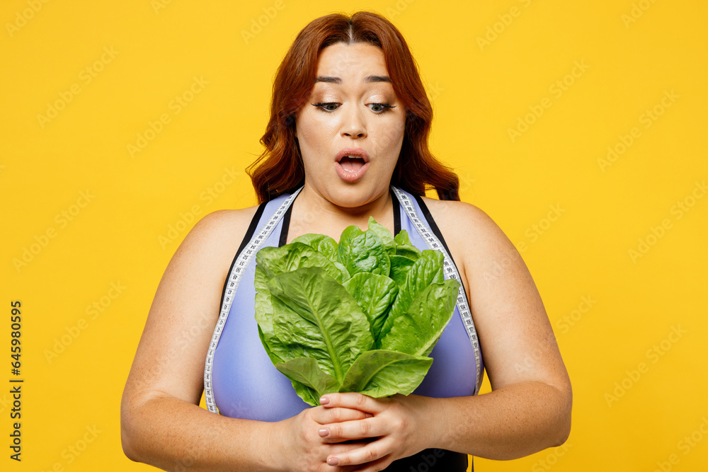 Young shocked chubby overweight plus size big fat fit woman wearing blue top warm up train holding tape measure eat broccoli isolated on plain yellow background studio home gym. Workout sport concept. Young shocked chubby overweight plus size big fat fit woman wearing blue top warm up train holding tape measure eat broccoli isolated on plain yellow background studio home gym. Workout sport concept.