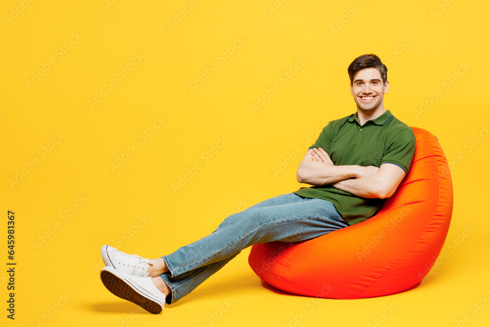 Full body side view young happy man he wears green t-shirt casual clothes sit in bag chair hold hands crossed folded look camera isolated on plain yellow background studio portrait. Lifestyle concept. Full body side view young happy man he wears green t-shirt casual clothes sit in bag chair hold hands crossed folded look camera isolated on plain yellow background studio portrait. Lifestyle concept.