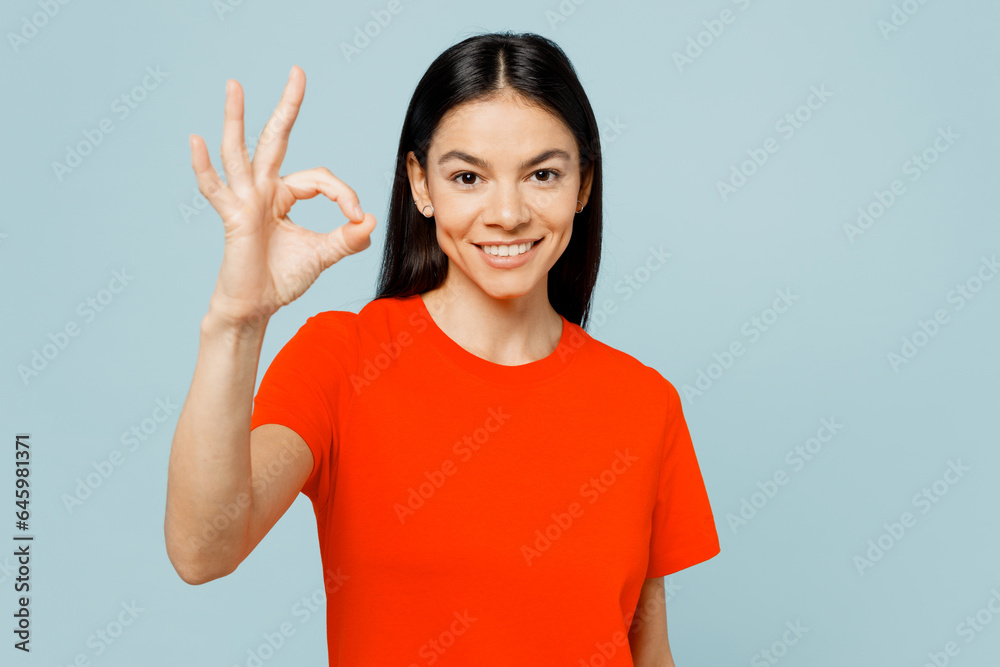 Young smiling happy cheerful fun latin woman she wear orange red t-shirt casual clothes showing okay ok gesture isolated on plain pastel light blue cyan background studio portrait. Lifestyle concept. Young smiling happy cheerful fun latin woman she wear orange red t-shirt casual clothes showing okay ok gesture isolated on plain pastel light blue cyan background studio portrait. Lifestyle concept.
