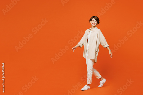 Full body side profile view young happy smiling caucasian woman she wear beige shirt casual clothes looking camera walk go isolated on plain orange red background studio portrait. Lifestyle concept.