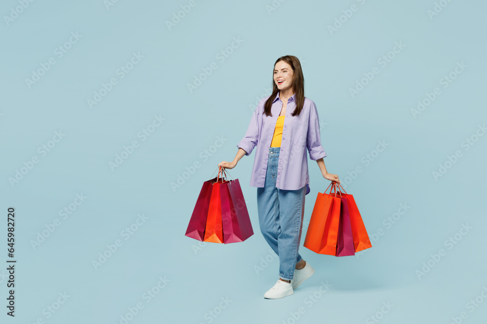 Full body young woman wearing purple shirt yellow t-shirt casual clothes hold paper package bags after shopping isolated on plain light pastel blue cyan background. Black Friday sale buy day concept.
