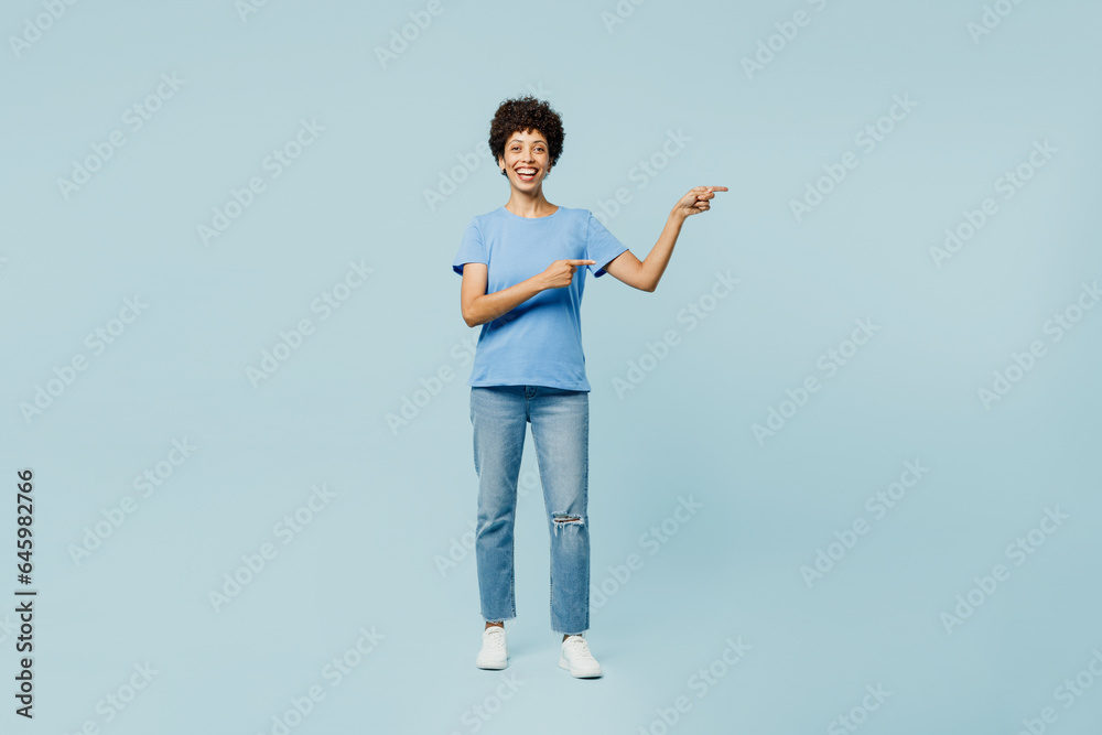 Full body young woman of African American ethnicity wears t-shirt casual clothes point index finger aside indicate on workspace area copy space mock up isolated on plain pastel blue cyan background. Full body young woman of African American ethnicity wears t-shirt casual clothes point index finger aside indicate on workspace area copy space mock up isolated on plain pastel blue cyan background.